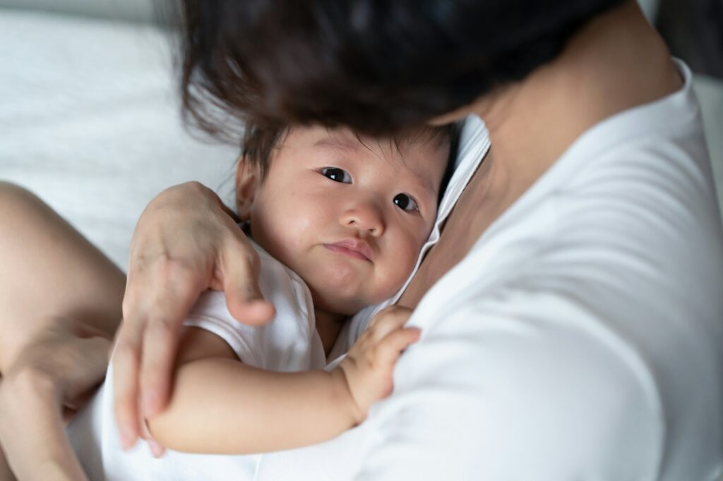 Asian mother holding crying baby in her arms and soothing her little daughter on the bed.